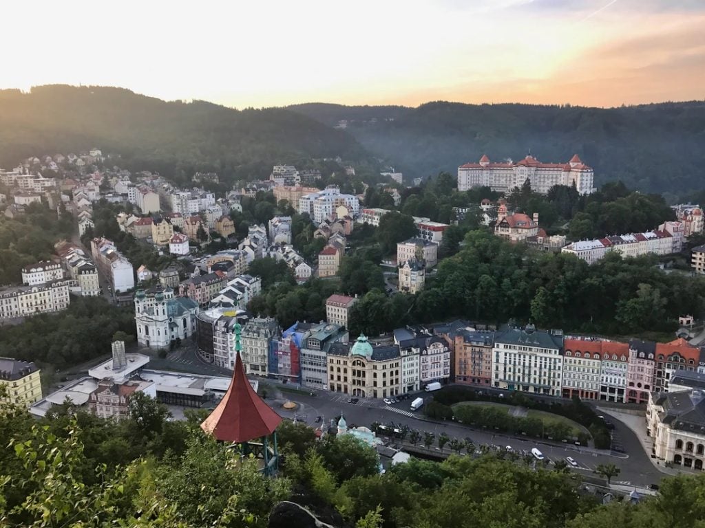 A city of pastel-colored crenellated buildings curving along the streets and surrounded by green trees. On the highest hill is what looks like a castle (or maybe a fancy hotel?), a white building with an orange roof.