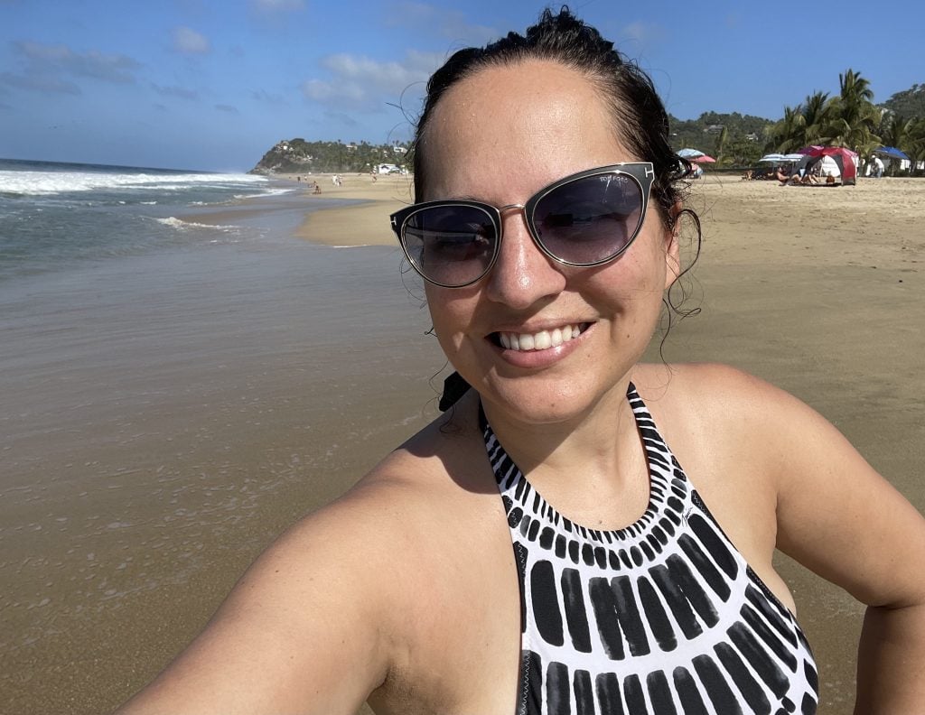 Kate taking a smiling selfie in front of the beach in San Pancho. She wears black sunglasses with gold rims and a black and white patterned bathing suit.