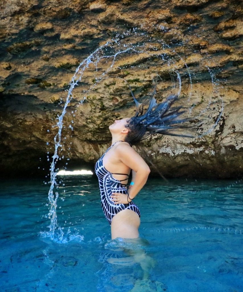 Kate stands facing sideways in bright blue water in a cave, in a black and white patterned bathing suit, hands on hips, and she whips her wet hair backwards, making her hair fling outward in a star pattern as it brings water droplets in an arc above her head.