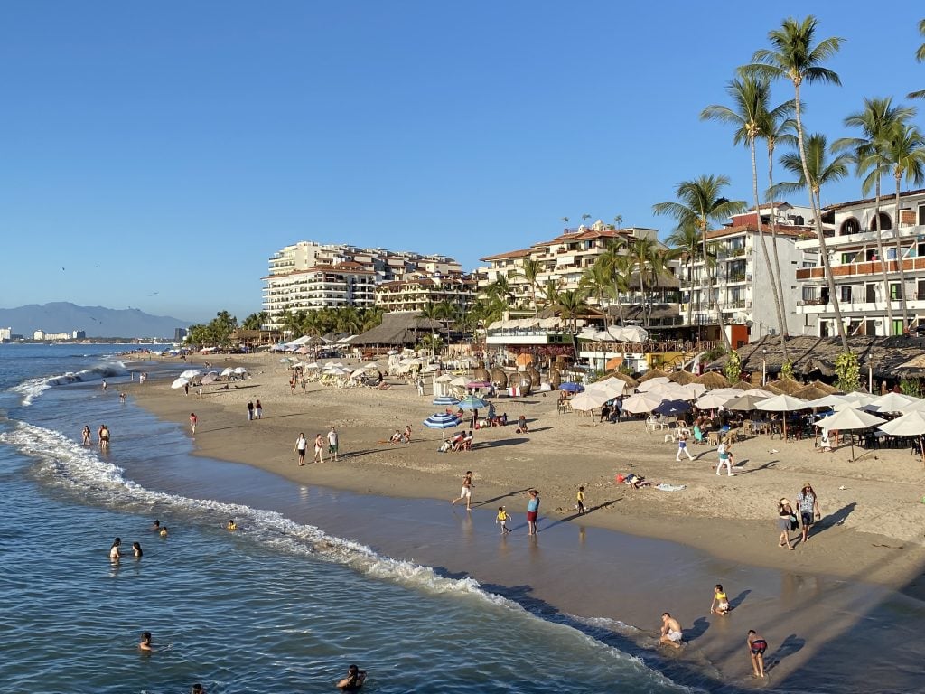 Los Muertos Beach in Puerto Vallarta: tall white buildings on land, lots of thatched roof beach bars and restaurants, and several people swimming in the surf, bright light just before sunset.