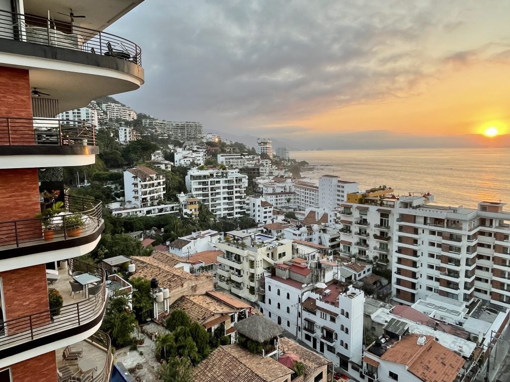 A view of hundreds of white high-rise buildings with balconies, ending with the water and a purple and orange sunset. The view is taken from another high rise that happens to be a cocktail bar.