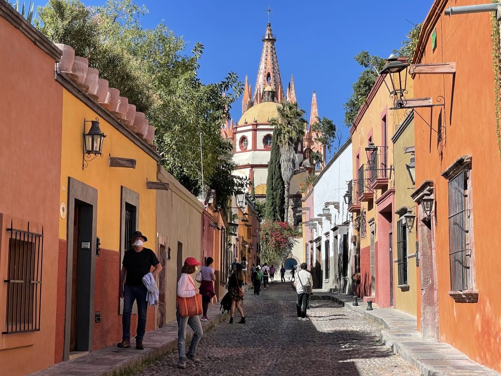 A cobblestoned street in San Miguel. On each side are squat buildings painted in warm shades of orange, brown, red, and mustard. In the center is San Miguel's cathedral in the background, with a golden dome, and behind it, several pink spikes.