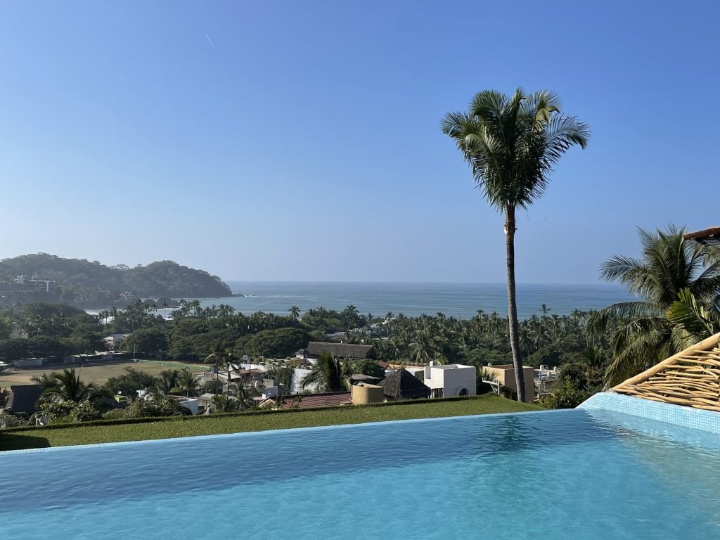 An infinity pool, and in the distance, a view of Sayulita -- lots of green palm trees, including one very tall palm tree, and a soccer field in the distance, leading to the beach and the ocean.