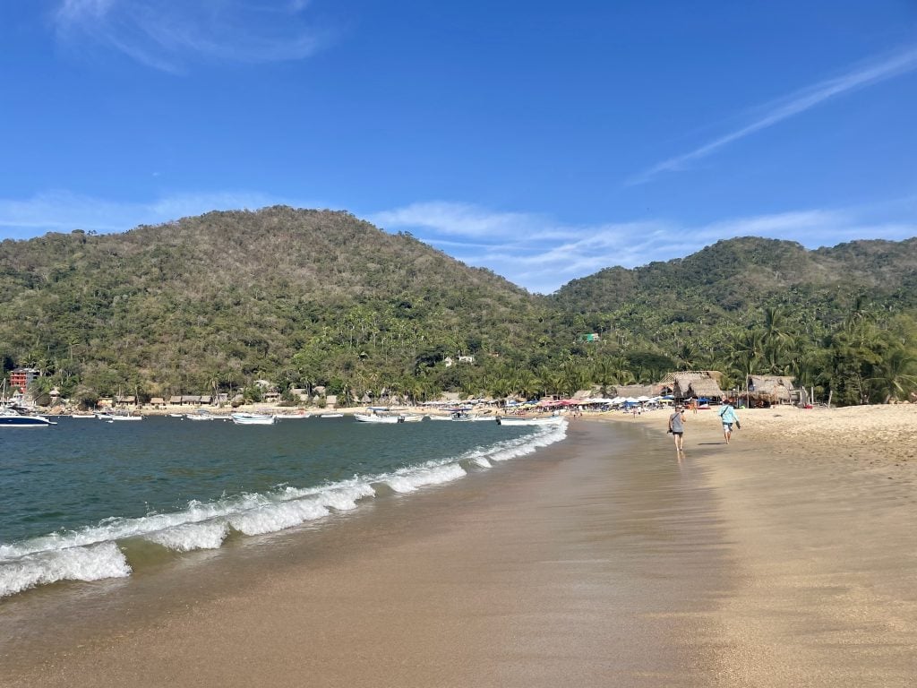 Yelapa beach, an almost empty golden sand beach with bright blue waves. In the distance you see several thatched roof beach bars and restaurants. Behind it, bright green mountains.