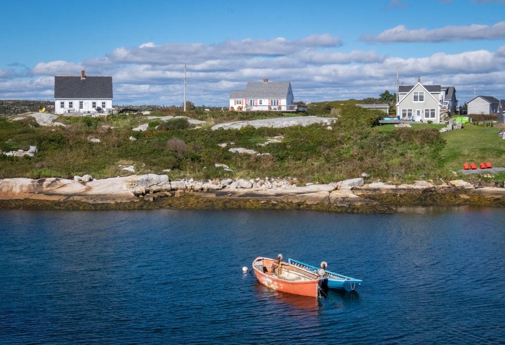 Two small red and blue rowboats in front of a grassy cliff with three cottages perched on top.