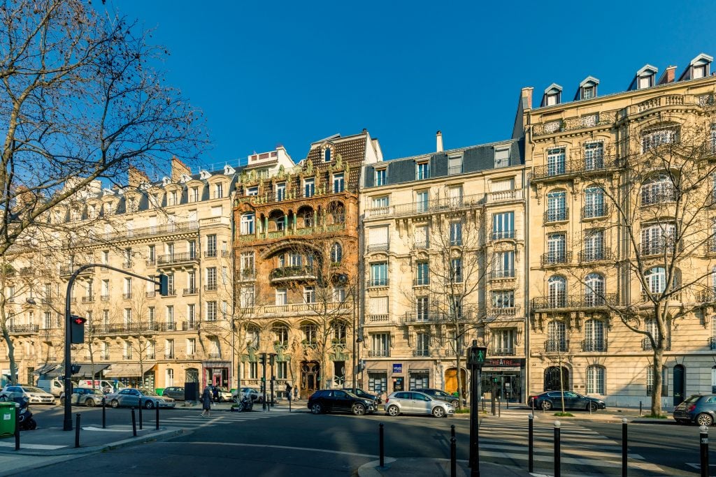 A normal-looking street of gray buildings in Paris, but in the middle is a wild gray-and-green building with lots of curves and curlicues in the architecture.