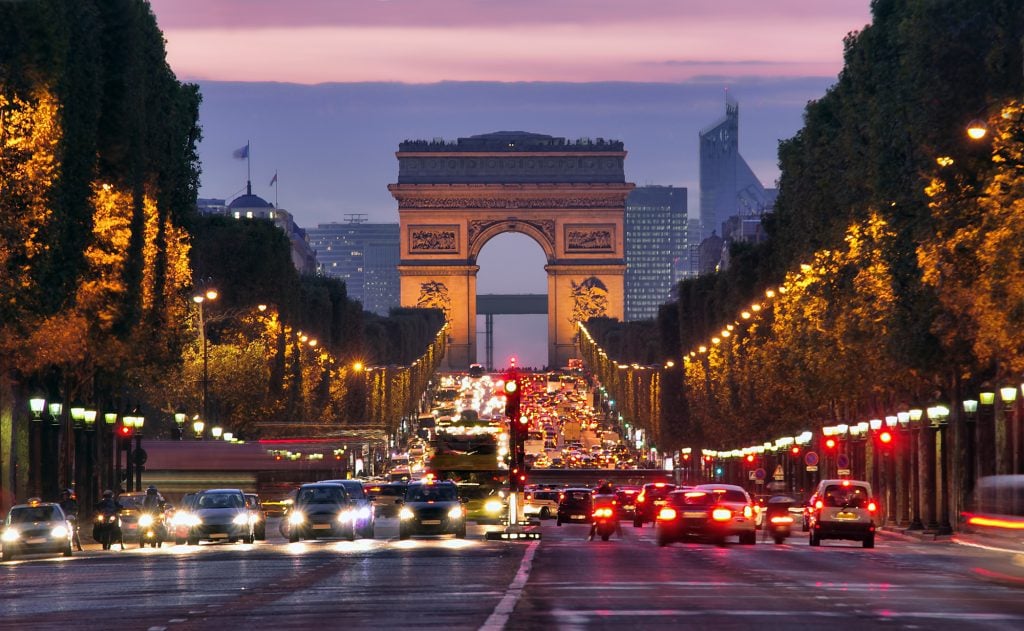 The Arc de Triumph at sunset, shining in evening light, and in front of it, the several lanes of the Champs-Elysees, with lots of traffic and bright car lights.