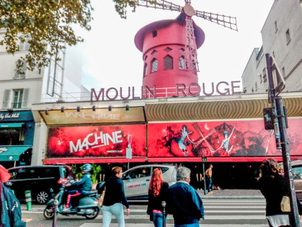 A street level view of the Moulin Rouge theater in Paris, a big red windmill on top.