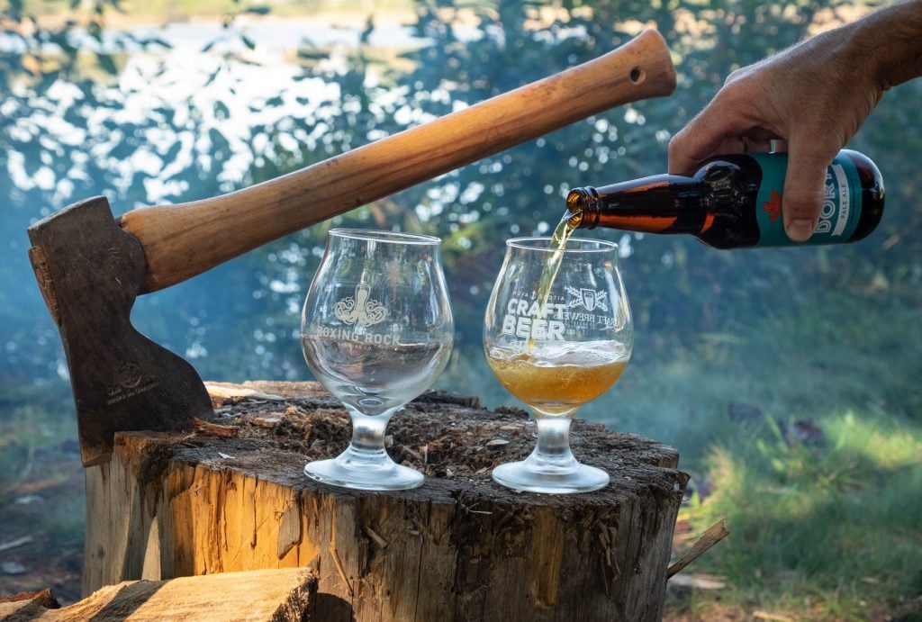 On a wooden stump, an axe blade perched in it, and two beer glasses, a hand pouring beer into one glass.