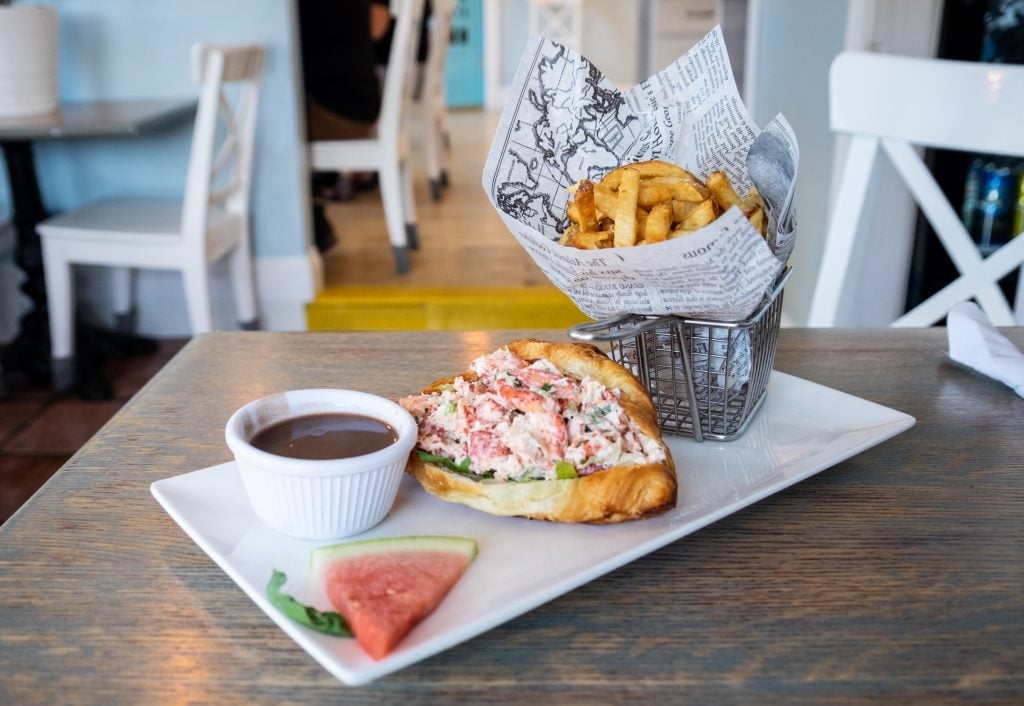 A lobster roll made out of a buttery croissant instead of a roll, next to a container of fries wrapped in faux newsprint, and a ramekin of gravy.