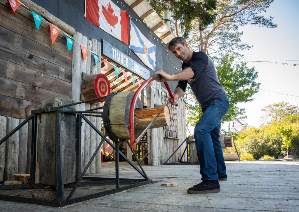 Darren: tall, thin, dark hair, cutting a log with a saw on the timber lounge stage.