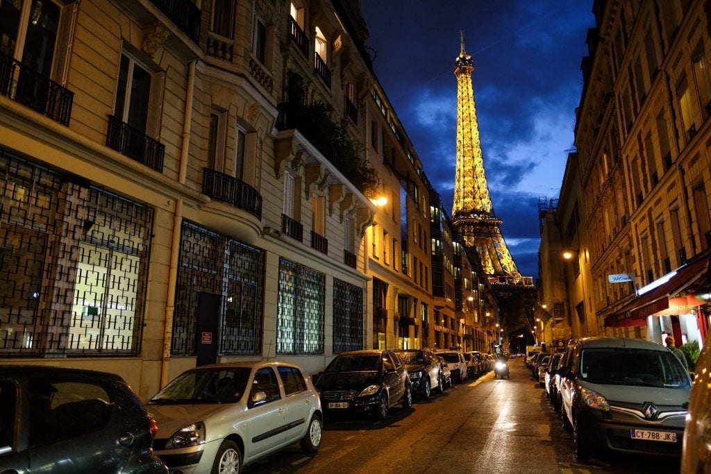 Paris at night, on a long street of gray buildings with some cafes and a motorcycle barreling down the street. In the background is the Eiffel Tower, lit up in bright gold against a dark blue cloudy sky.