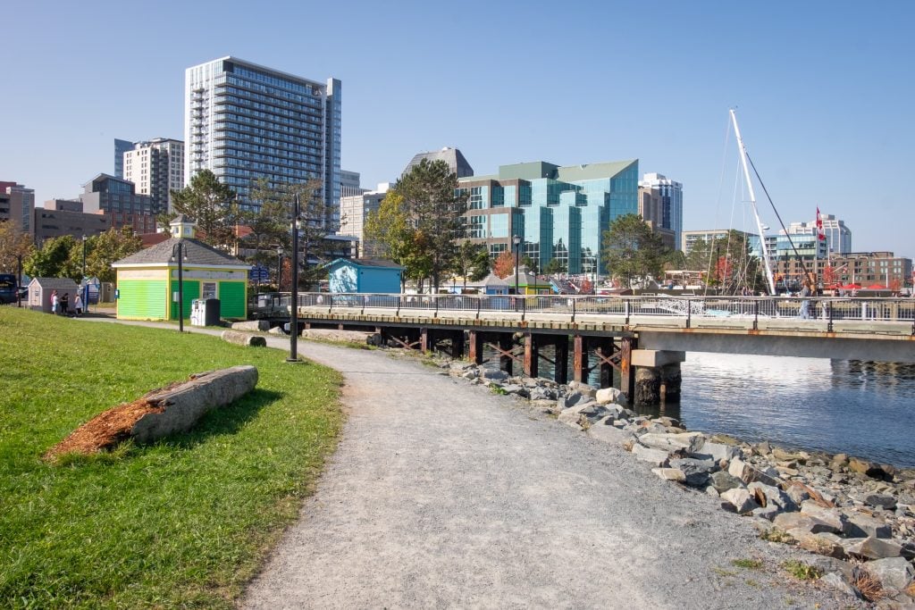 A path running alongside the water; you see several tall skyscrapers.