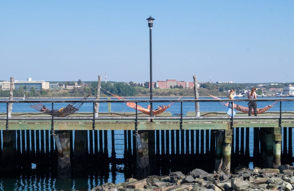 A wooden pathway over the harbor with three strung up hammocks, a person in each of them.