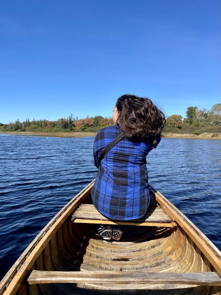 Kate sitting at the front of a canoe, the shot from behind. The sky and pond are bright blue and Kate runs a hand through her long dark hair.