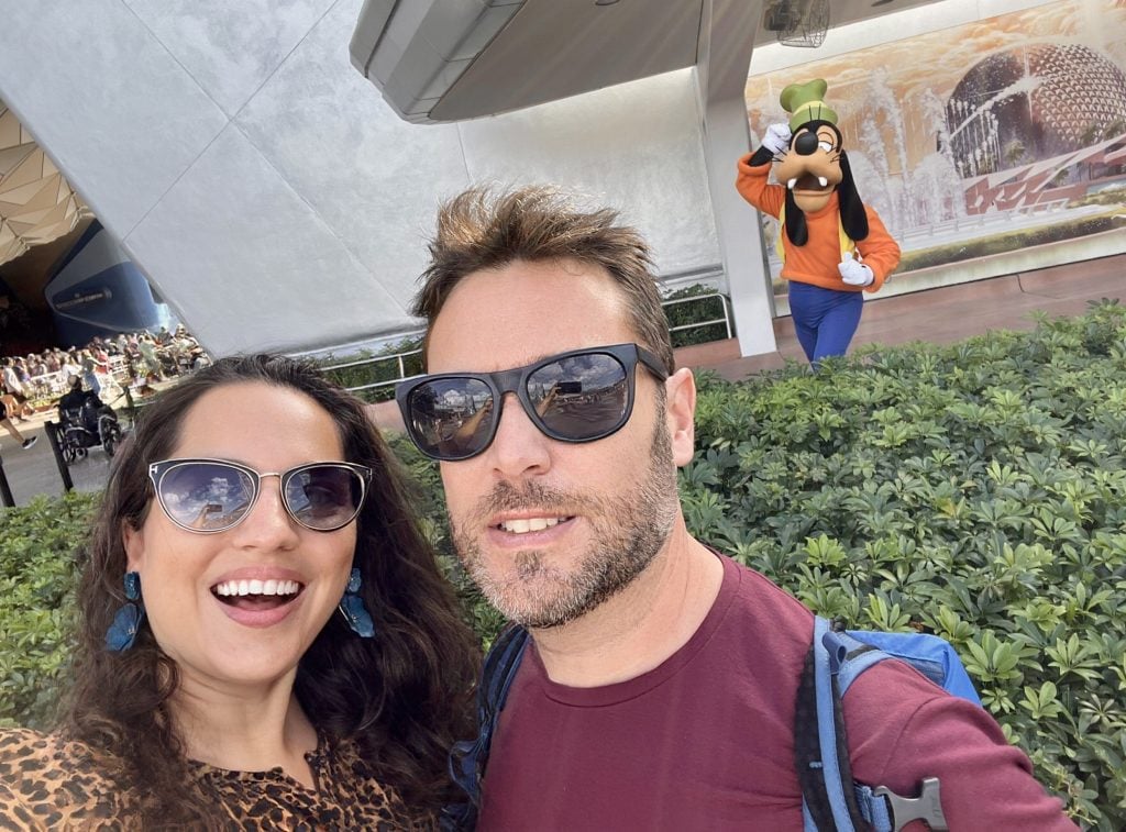 Kate and Charlie taking a selfie in sunglasses in front of a large hedge, and behind the hedge is Goofy.