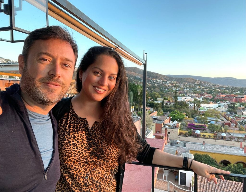 Kate and Charlie smiling and taking a selfie at a rooftop bar; behind them are the rolling hills of San Miguel, dotted with houses.