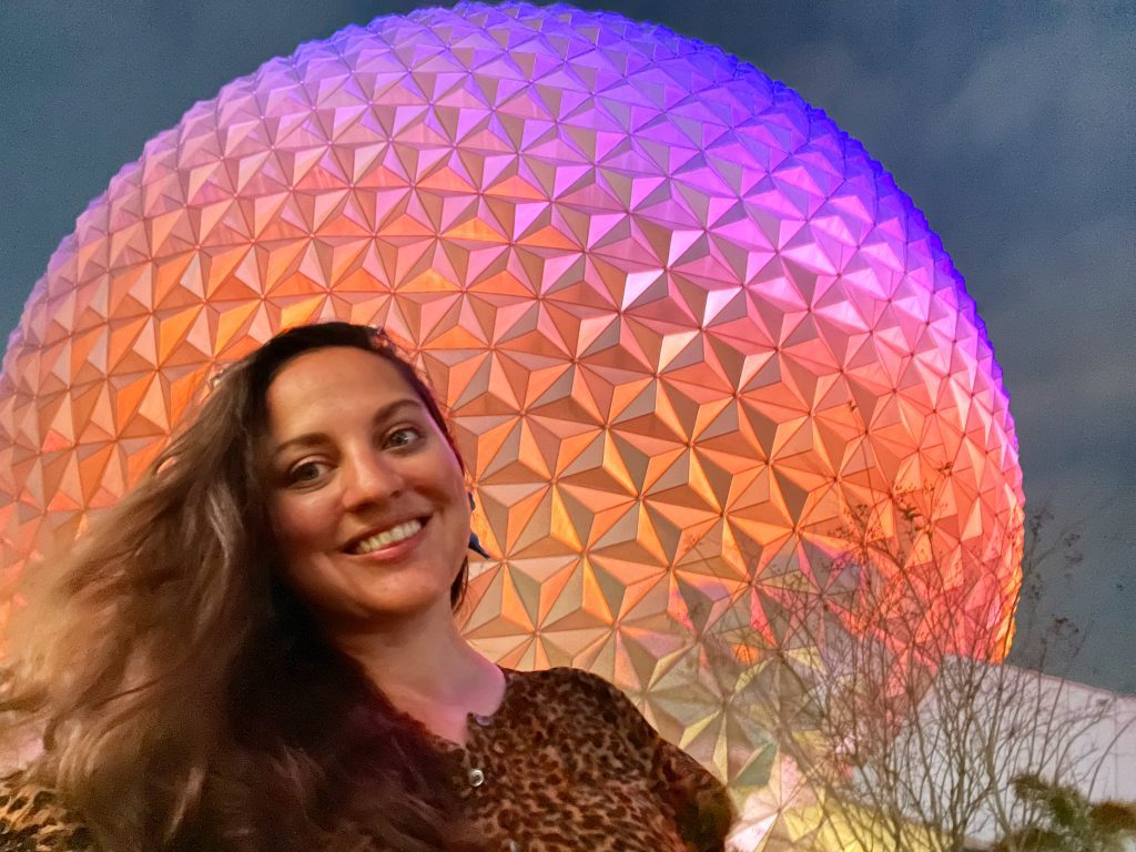 Kate taking a smiling selfie in front of Spaceship Earth, the dome at Epcot, lit up in purple, pink, orange, and yellow.