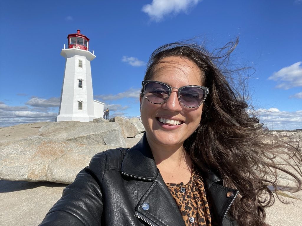 Kate taking a smiling selfie in a leopard-print shirt and black leather jacket with sunglasses on. Behind her is the white Peggy's Cove lighthouse perched on the rocks.