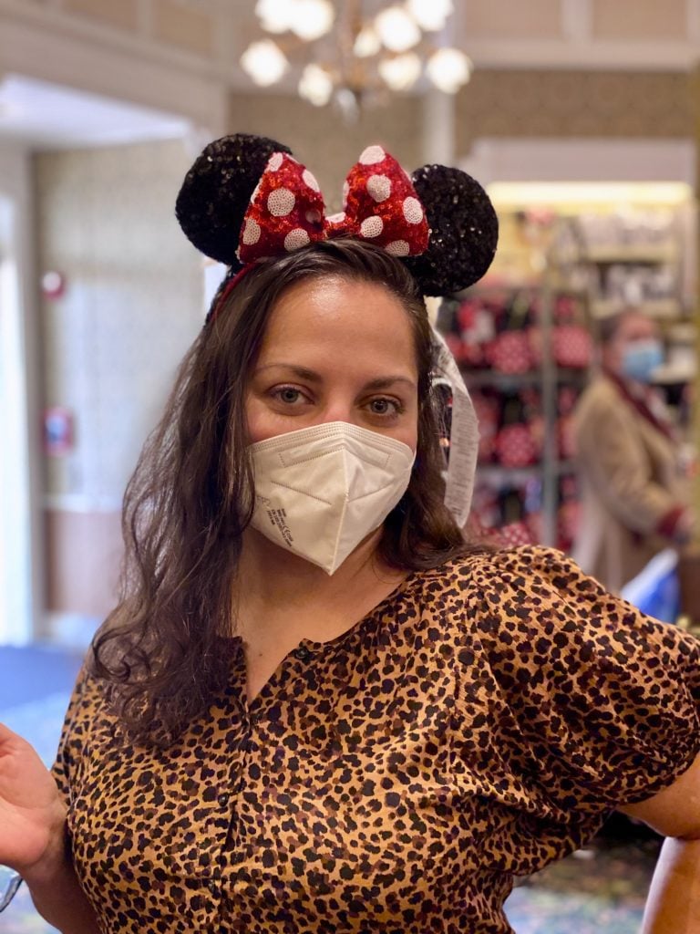 Kate smiling in a leopard-print blouse and a white face mask, with a pair of sparkly Minnie Mouse ears on her head.