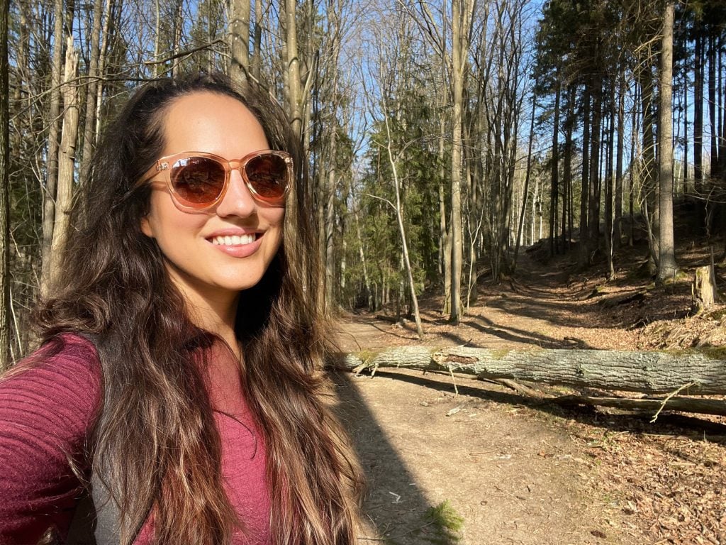 Kate wears sunglasses, her hair long, and stands in front of a wooded trail.