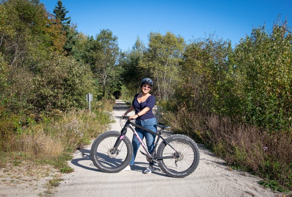 Kate smiling, posing while holding her bike on a path going through the woods.