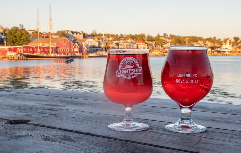 Two glasses of bright red cranberry cider with the Lunenburg waterfront lit up in sunset light in the background.