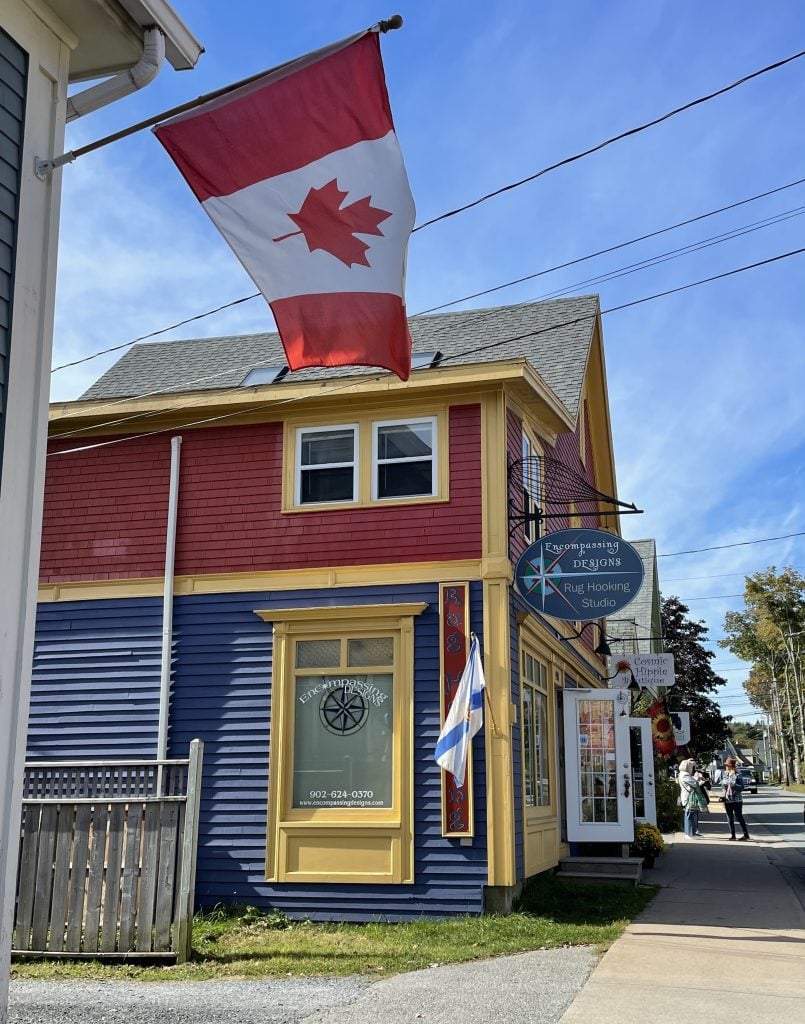 A cottage painted bright primary colors with a sign reading "Rug Hooking Studio," a Canadian flag flying overhead.