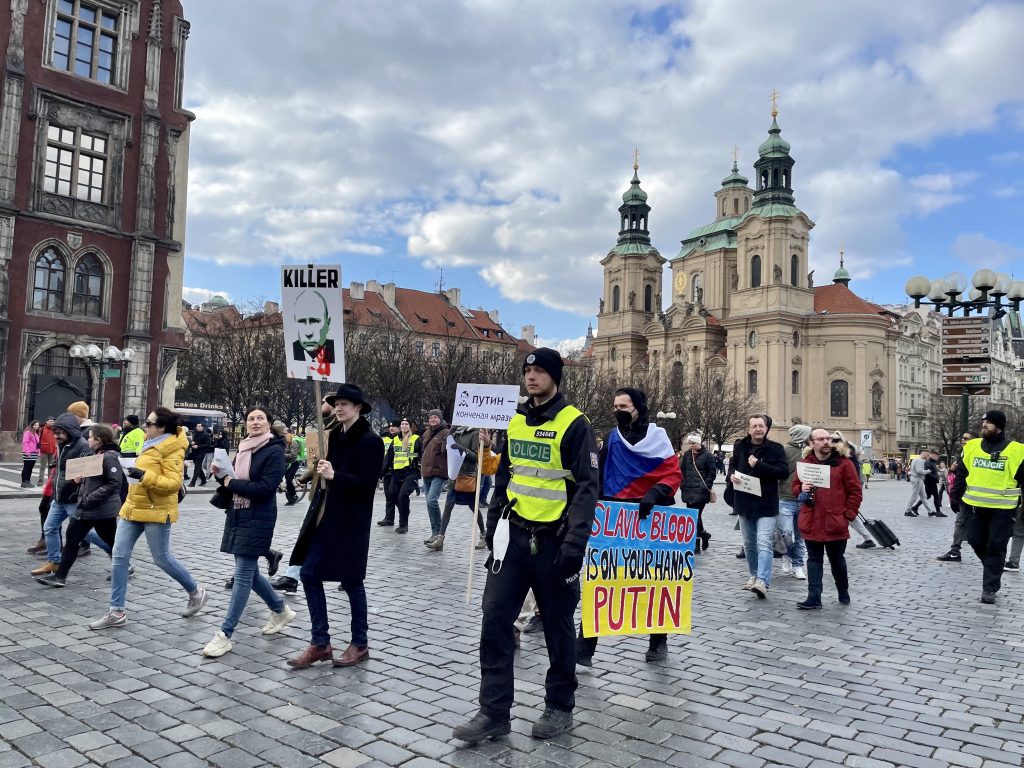 People marching in Prague's Old Town square, in front of a big gray church, to protest the war in Ukraine. One sign has Putin's face and says Killer; another says SLAVIC BLOOD IS ON YOUR HANDS PUTIN.