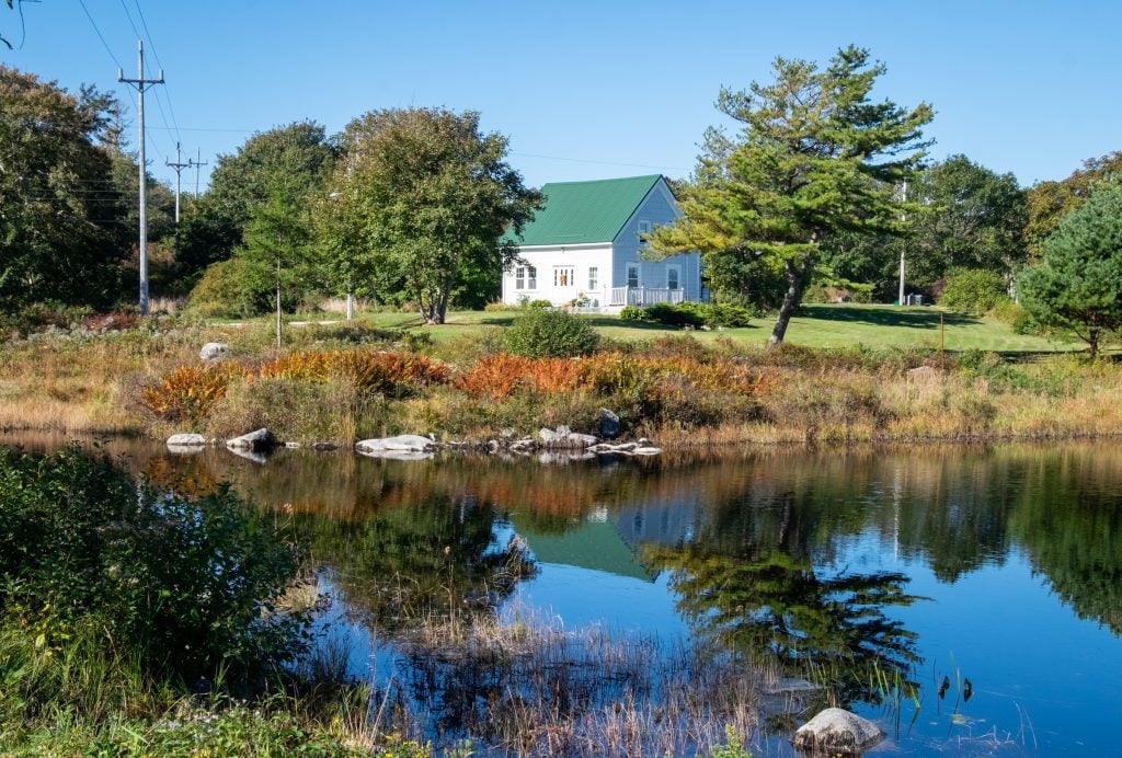 A small white cottage perched on the edge of a calm pond, surrounded by trees.