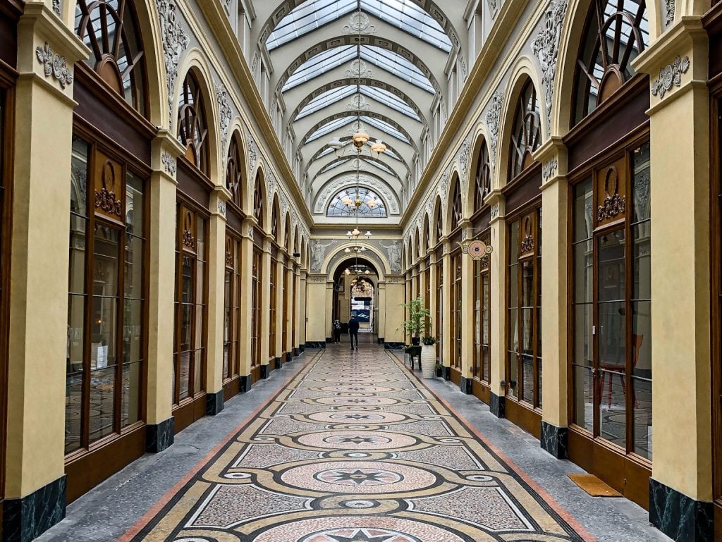 A view down a hallway with a mosaic floor and arched glass window ceiling, with windows to various shops along the way