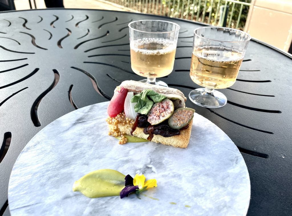 A small piece of bread topped with pate, mustard, and caper berries, next to two glasses of sparkling wine.