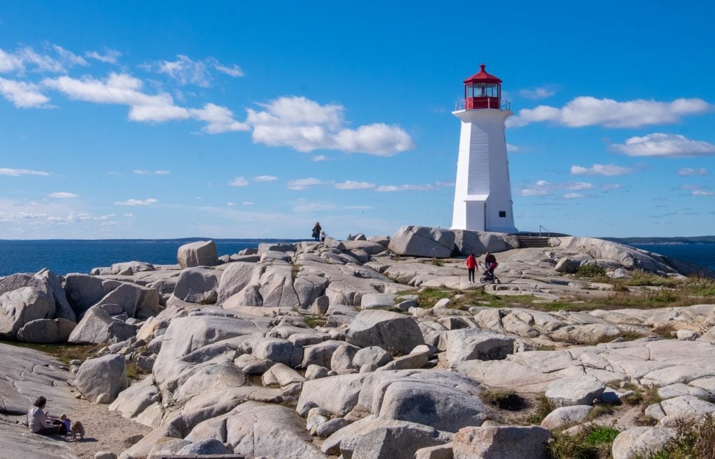 The big white Peggy's Cove lighthouse topped with a red top, on a large rocky expanse in front of the ocean.