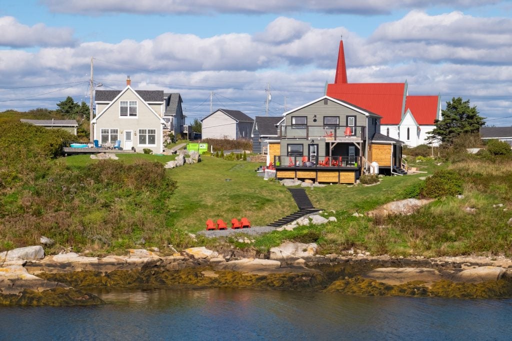 A close-up view of two cottages and a church on the edge of the sea, with four red Adirondack chairs by the water.