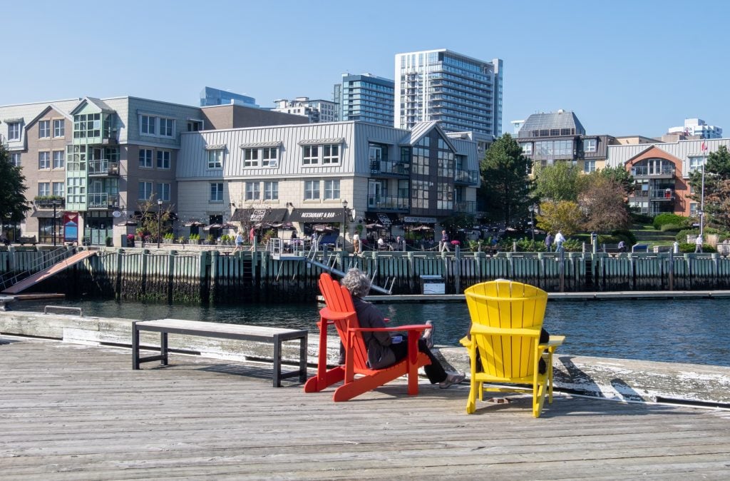 Two people sitting in red and yellow Adirondack chairs overlooking the skyscrapers on the Halifax waterfront.