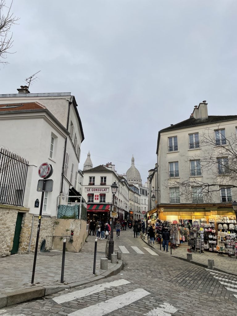 An empty Parisian street on a cloudy day, with three gray buildings in the background. A yellow awning over a cafe is on the corner of one of them.