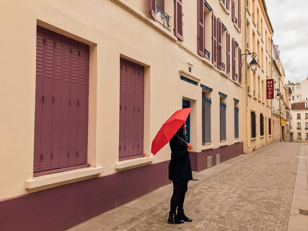 A woman dressed in black holding a bright red umbrella in front of a cream colored building on Rue Paulin in Paris