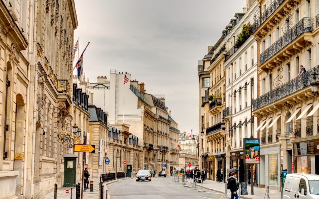 The bend of a Parisian street, with a few cars on the road and people on balconies