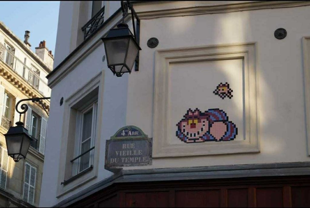 The corner of a building that says Rue Vielle du Temple with two streetlights hanging and a a painting of the cheshire cat