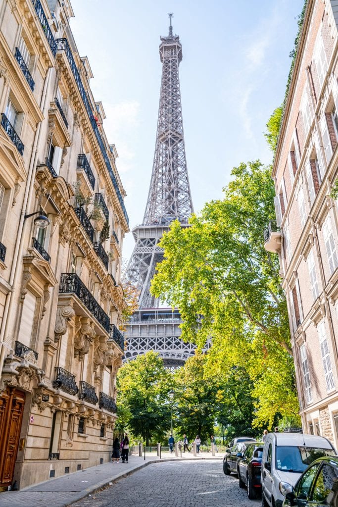 View of the Eiffel tower down a Parisian street with cars parked on the righthand side and bright green trees in front of the tower