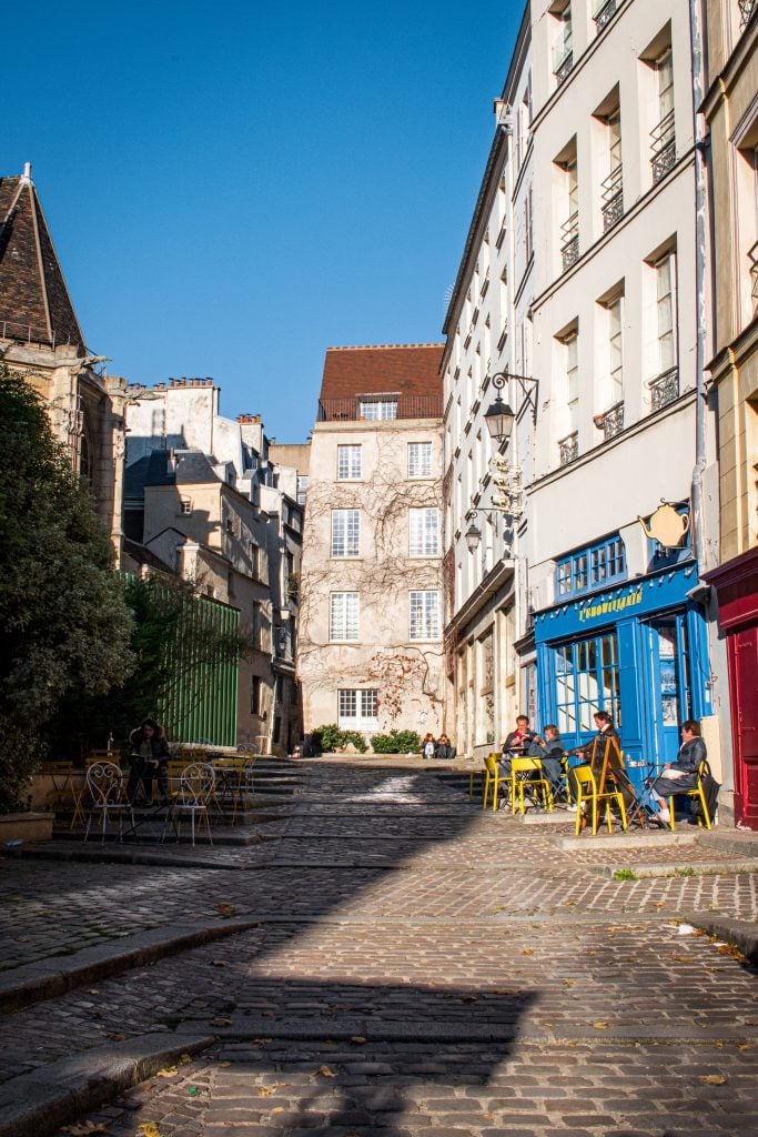 A street view of the uphill cobblestoned Rue des Barres in Paris, with a small blue cafe with yellow chairs on the right