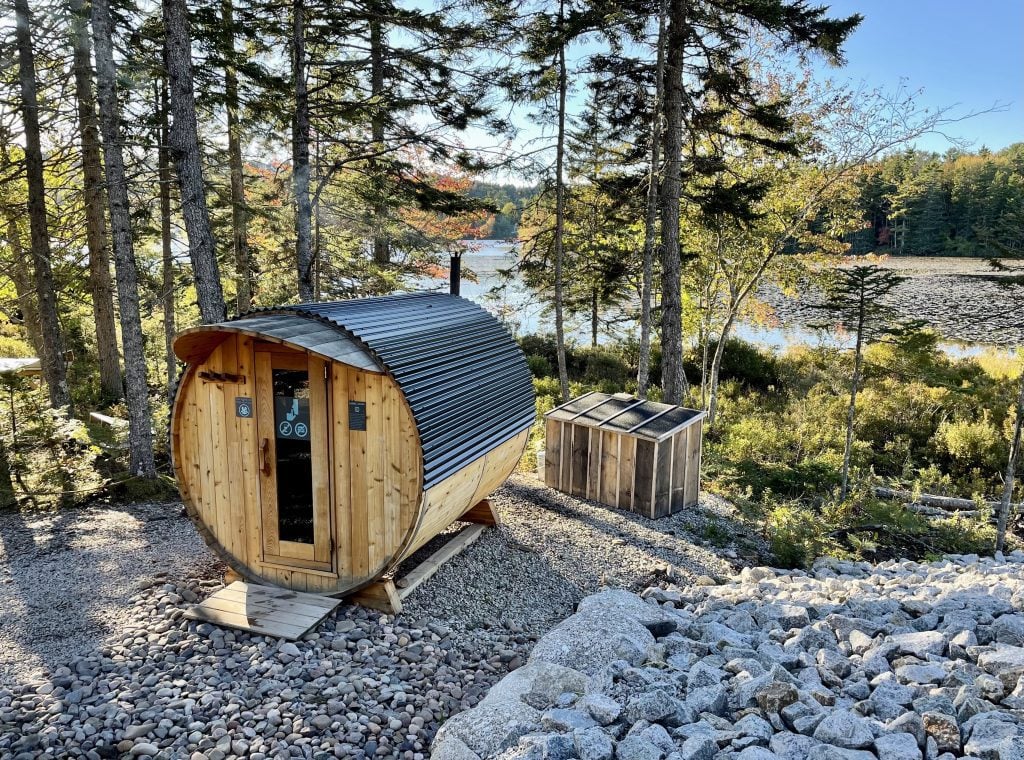 A tiny round wooden house that is actually a small sauna sitting in the forest by the lake.