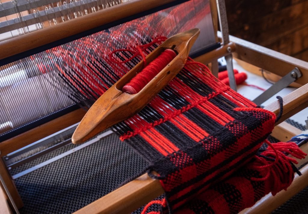 A red and black scarf being woven by hand with the thread sitting in a wooden boat you pass back and forth underneath the loom.
