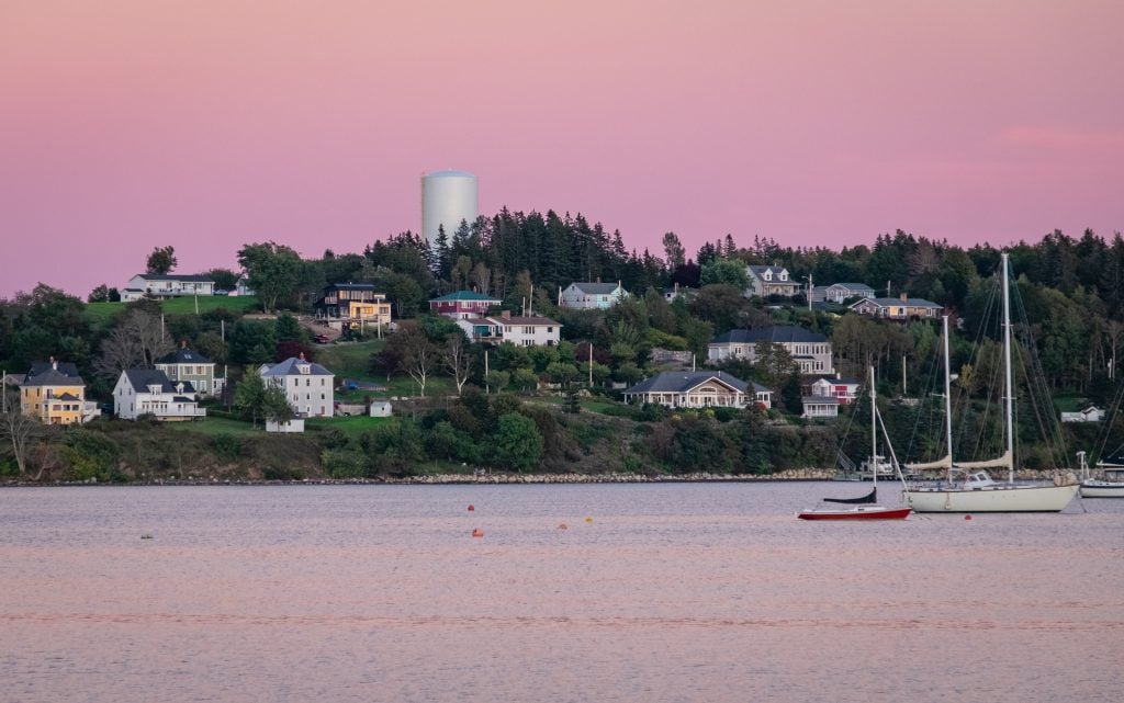 A hill in front of the harbor with several houses on it; above the sky is bright pink for sunset.
