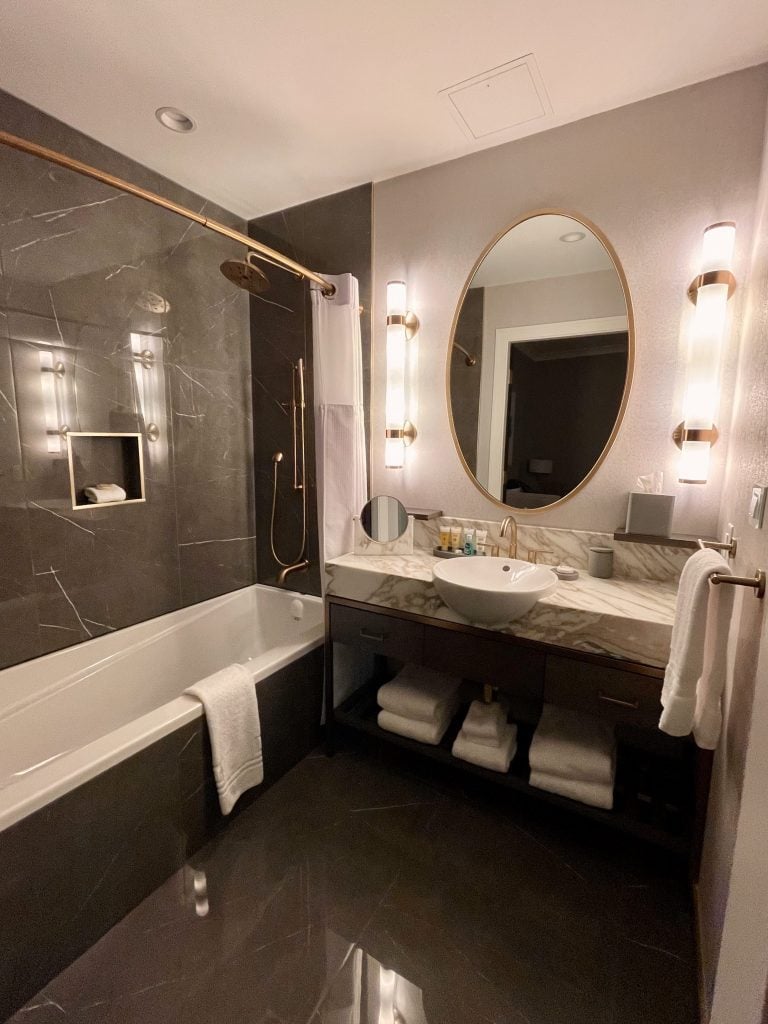 A luxury bathroom decorated in gray and gold, with a big oval gold mirror and a gray and white marble sink.