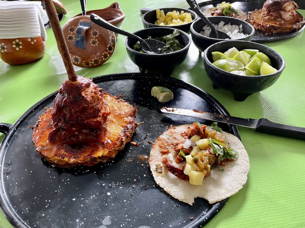 A plate with a spit of marinated cooked pork on it with a slice of pineapple as the bottom; next to it, a small taco filled with the pork, green salsa, onions, pineapple, and cilantro.