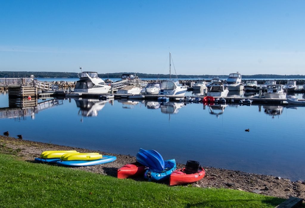 A calm blue harbor with several large boats docked at a pier. On land, some yellow, blue, and red kayaks lined up.