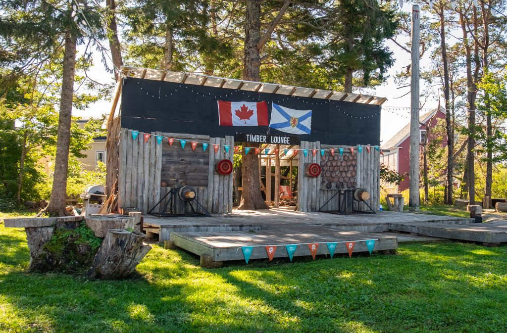 A large wooden stage that reads "Timber Lounge" and has the Canadian and Nova Scotian flags flying. Red and yellow flags hanging, and logs on stage ready to be cut.