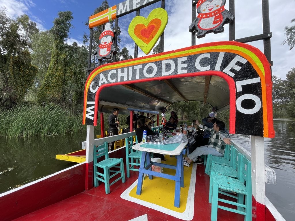 A large flat wooden boat reading "Mi Cachito De Cielo" on top, with people sitting at chairs around a table on board. Next to it is a mariachi band floating on another boat.