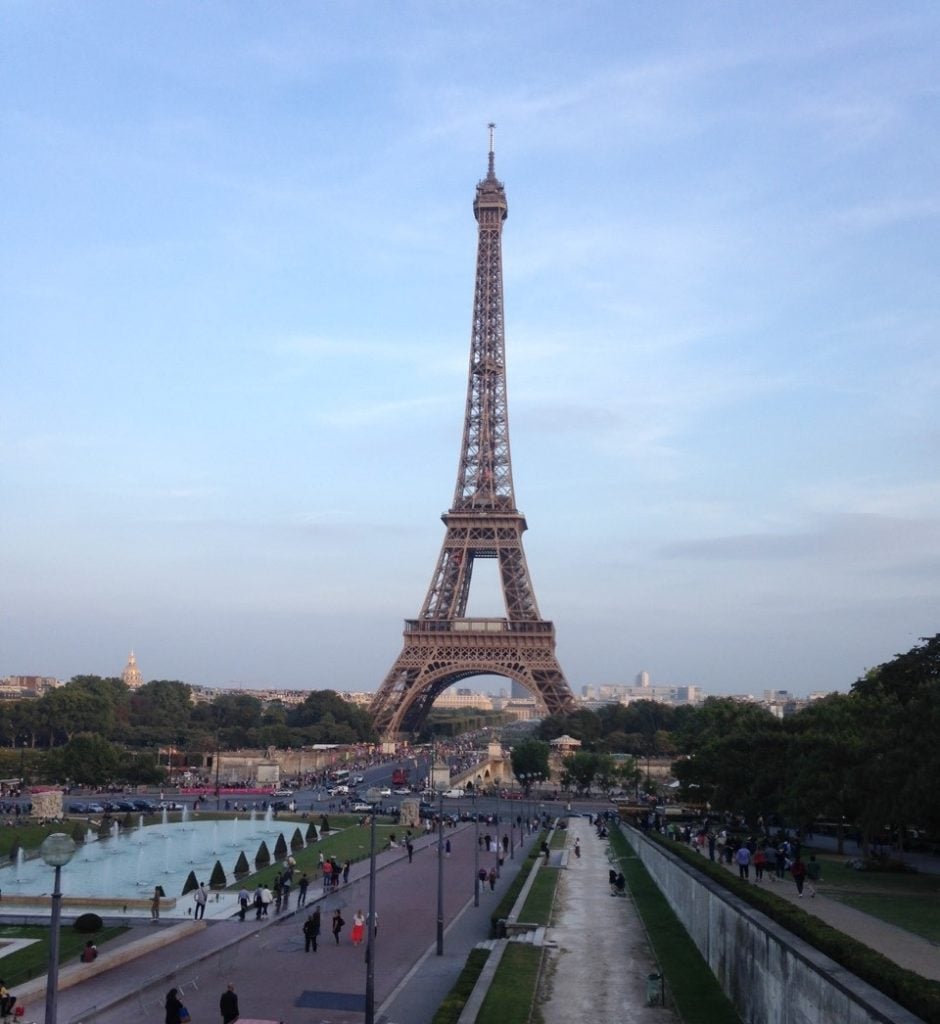 A view of the Eiffel tower from the lifted walkway along Avenue Albert. You can see the walkway to the right, a fountain to the left, and buildings behind the tower